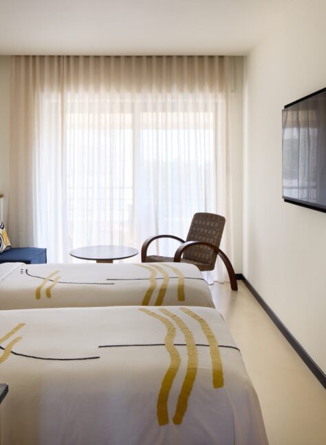 A modern hotel room with two neatly made beds featuring abstract patterned bedspreads. A large window with sheer curtains provides soft natural light. A cozy chair and small round table are near the window, and a wall-mounted TV is opposite the beds, creating a calm, inviting atmosphere.