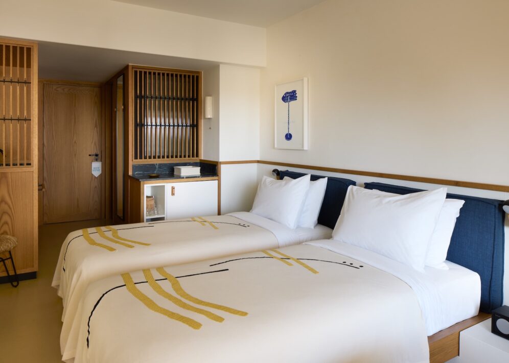 Modern hotel room with two neatly made single beds adorned with abstract-patterned blankets. Warm wooden accents, a small art piece, and soft lighting create a cozy atmosphere.