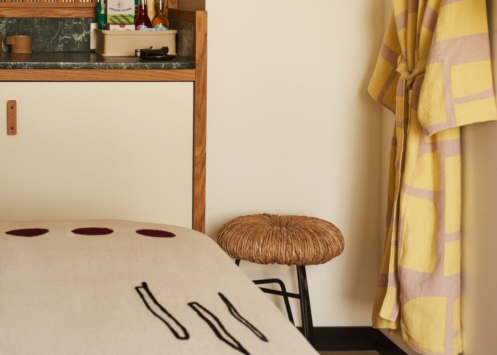 Cozy room corner with a yellow robe on a wall hook, woven stool, and a wooden countertop holding snacks and drinks. Warm, inviting atmosphere.