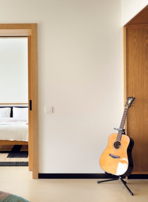 A serene room features a wooden door leading to a cozy bedroom with a white bed. A guitar stands beside a wood panel with artistic green designs.