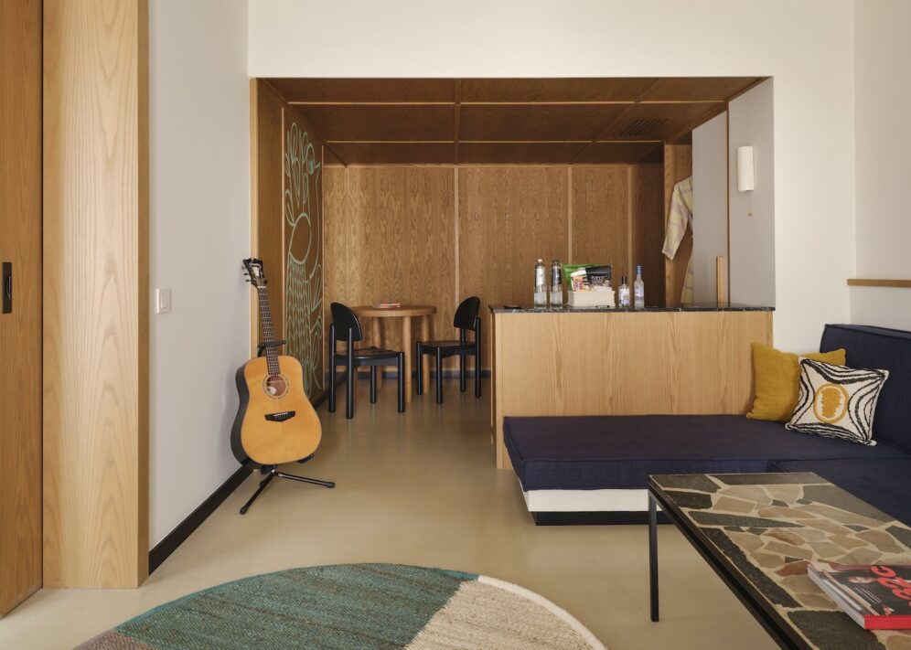 Cozy living space featuring a guitar on a stand, wood-paneled walls, blue sofa with vibrant cushions, mosaic coffee table, and dining area. Warm, inviting ambiance.