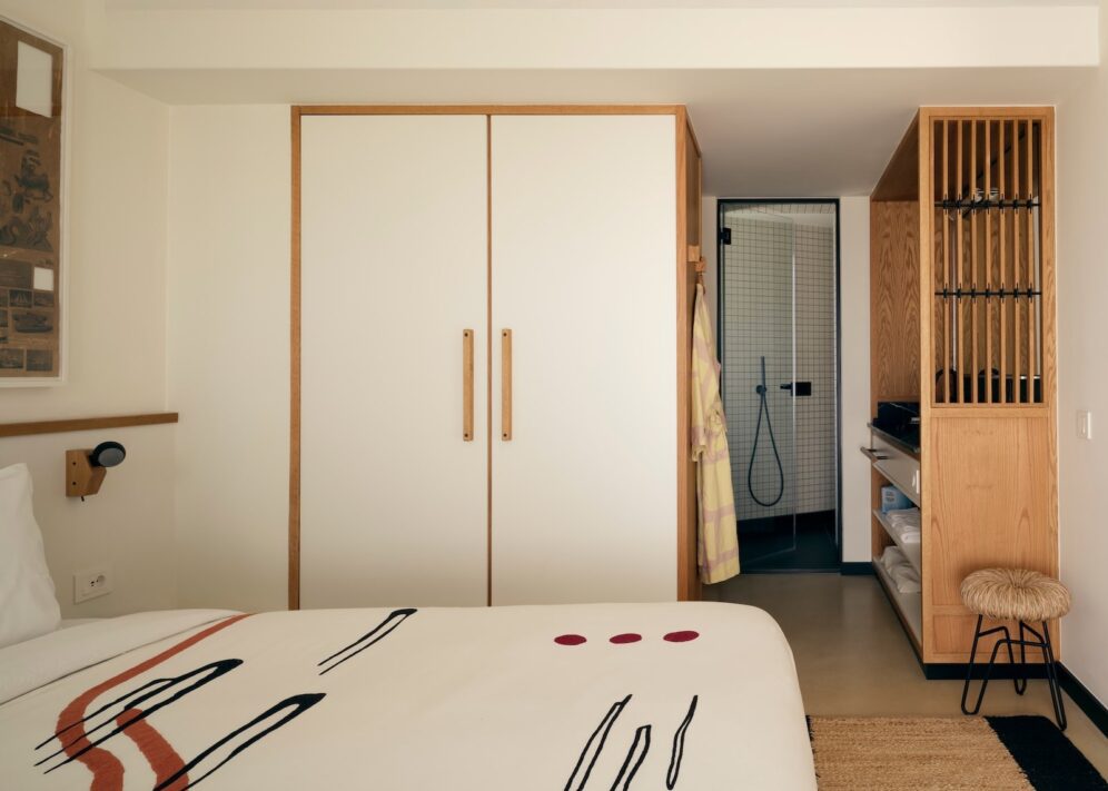 Cozy hotel room with a minimalist design. Features a bed with abstract-patterned bedding, a large wardrobe, open shelving, and a glimpse of a modern bathroom.