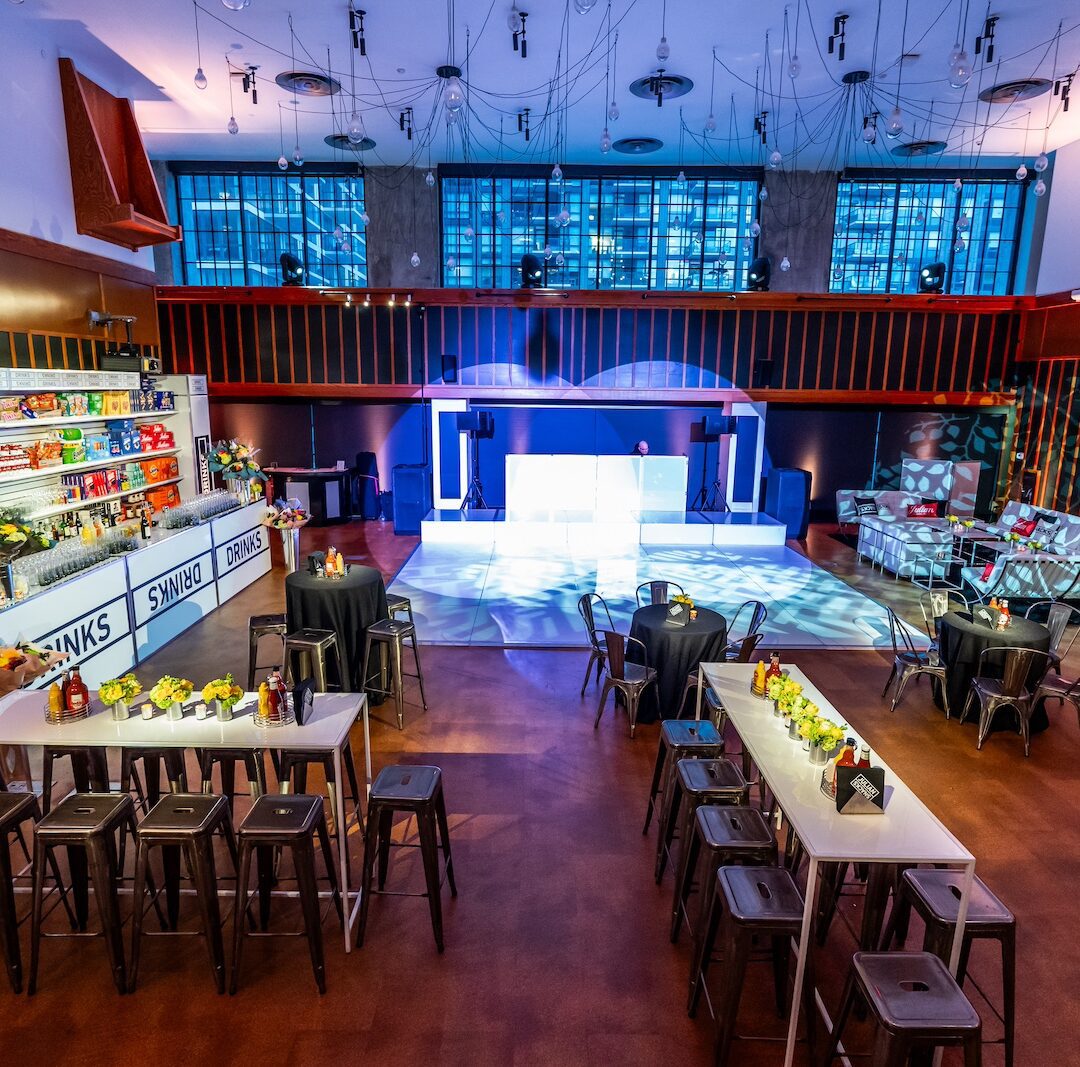 Spacious event venue with bar seating, high tables, and colorful lighting, featuring large windows and shelves stocked with snacks.
