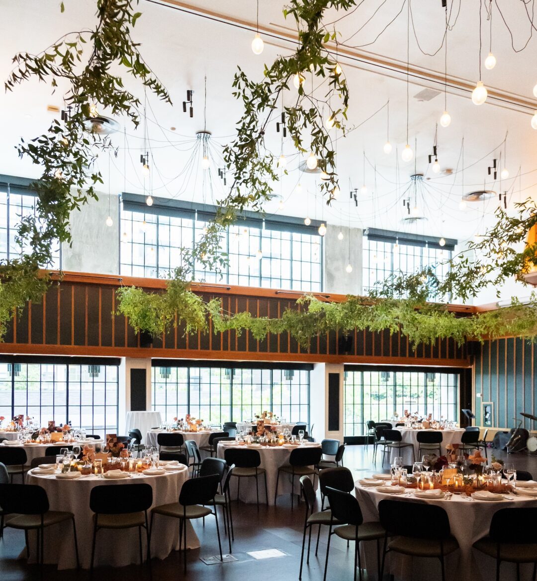 Spacious event hall with large windows, decorated with hanging greenery and round tables set for a formal gathering.