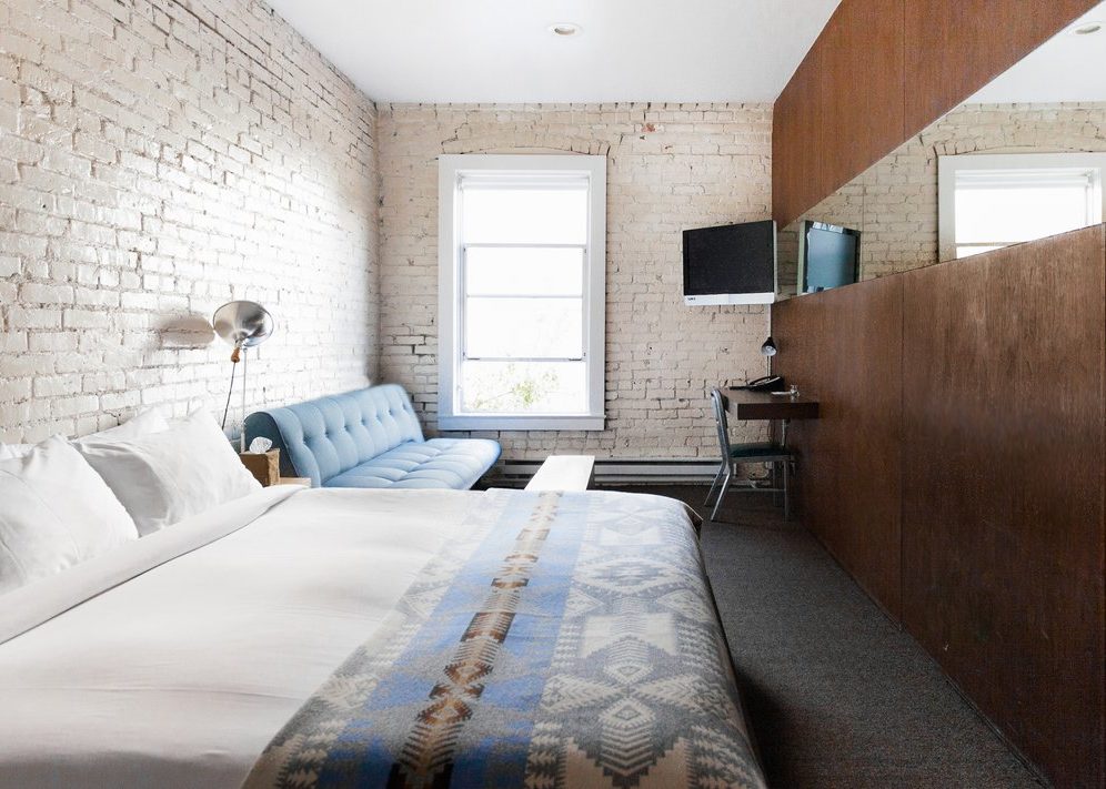 a room with exposed white brick walls is drenched in natural sunlight; a bed sits next to a long couch