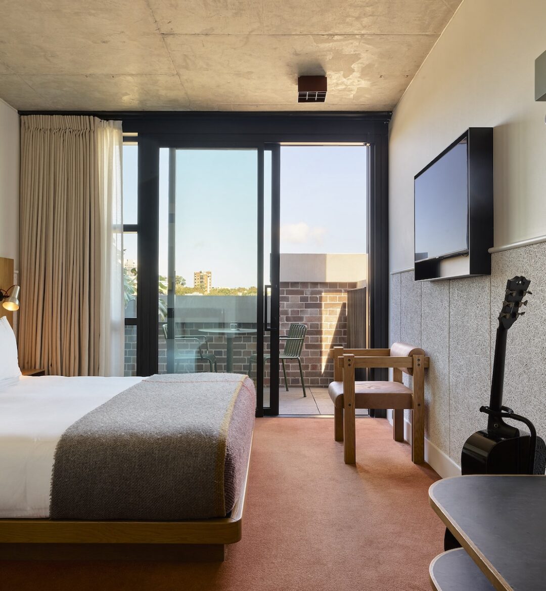 Modern hotel room with a double bed, TV, guitar, desk, and balcony with outdoor seating, featuring neutral tones and minimal decor.