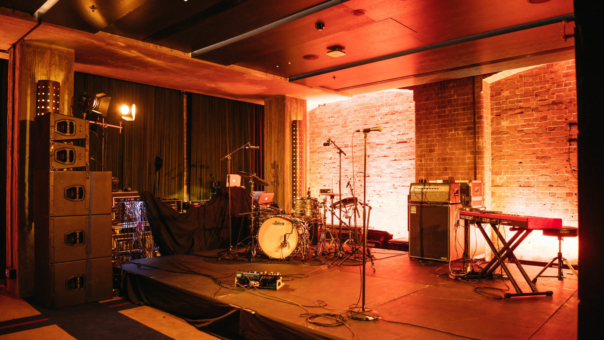 A small indoor stage with drum set, microphones, keyboard, speakers, and lighting, ready for a live music performance.