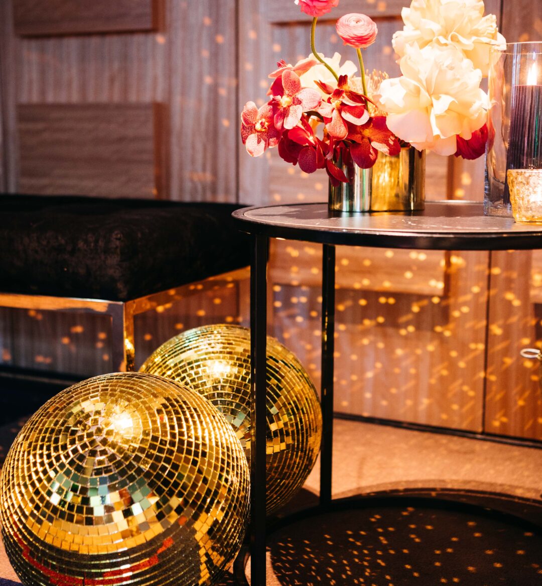 Gold disco balls rest on the floor next to a round table with a vase of red and white flowers and a small candle.