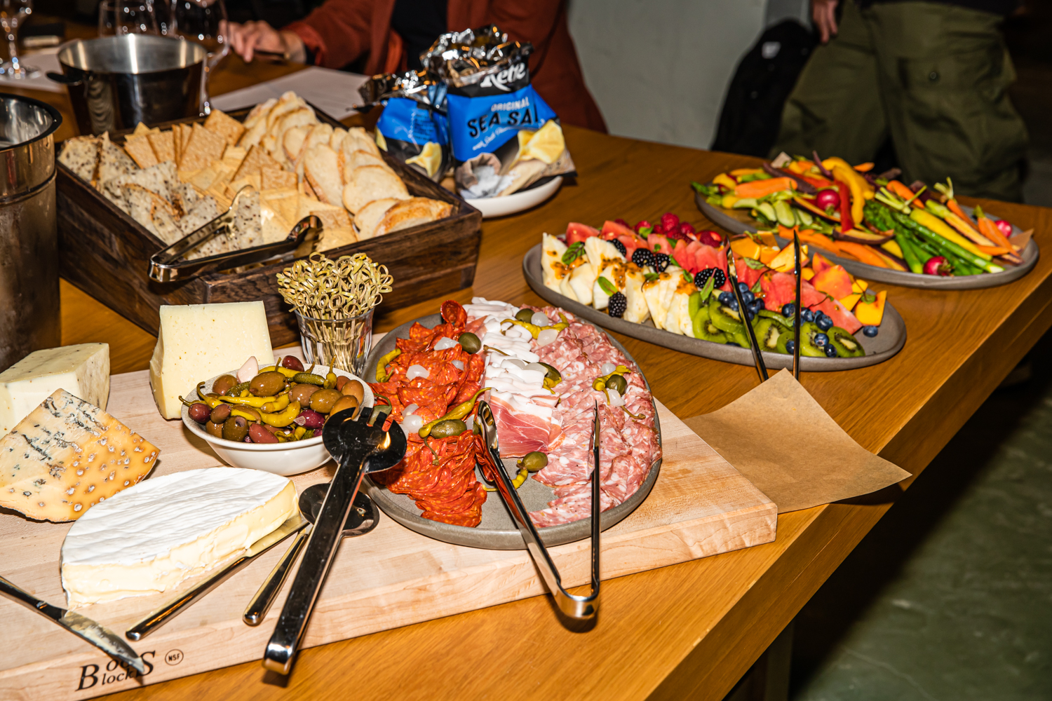 A wooden table with assorted cheeses, cured meats, olives, breads, chips, and colorful platters of fresh fruits and vegetables.