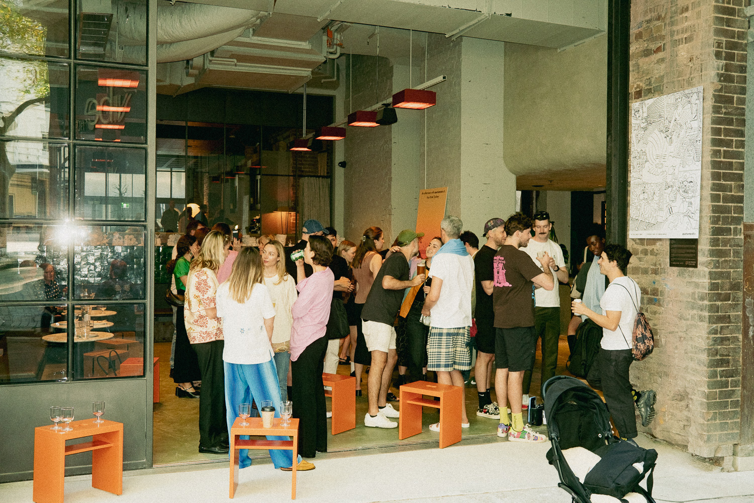 A group of people gather and socialize inside and outside a modern building, with tables, benches, and drinks visible.