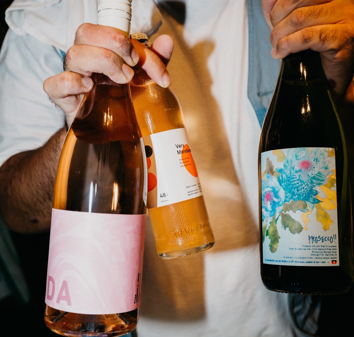 A person holds three bottles: one pink-labeled, one orange-labeled, and one with a floral design labeled “Prosecco!!”.