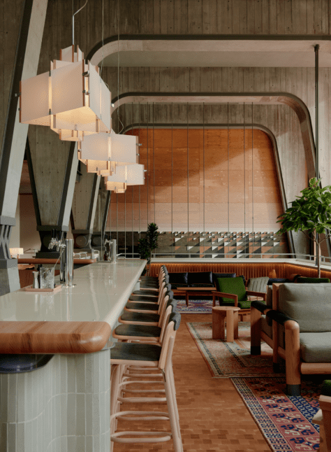 Modern cafe interior with a long bar, wooden chairs, pendant lights, and green lounge seating under large arched concrete beams.