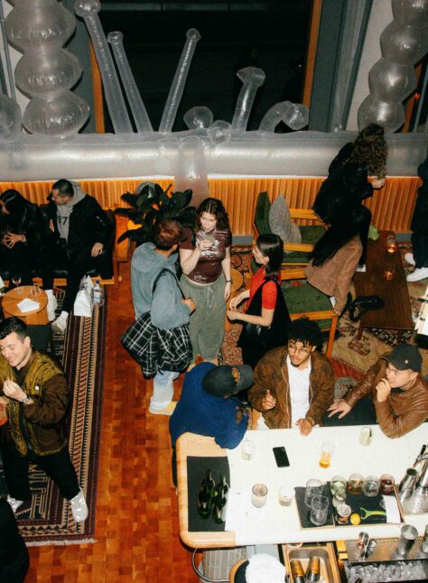 A group of people socializes in a lounge with drinks, couches, rugs, and warm lighting, viewed from above.
