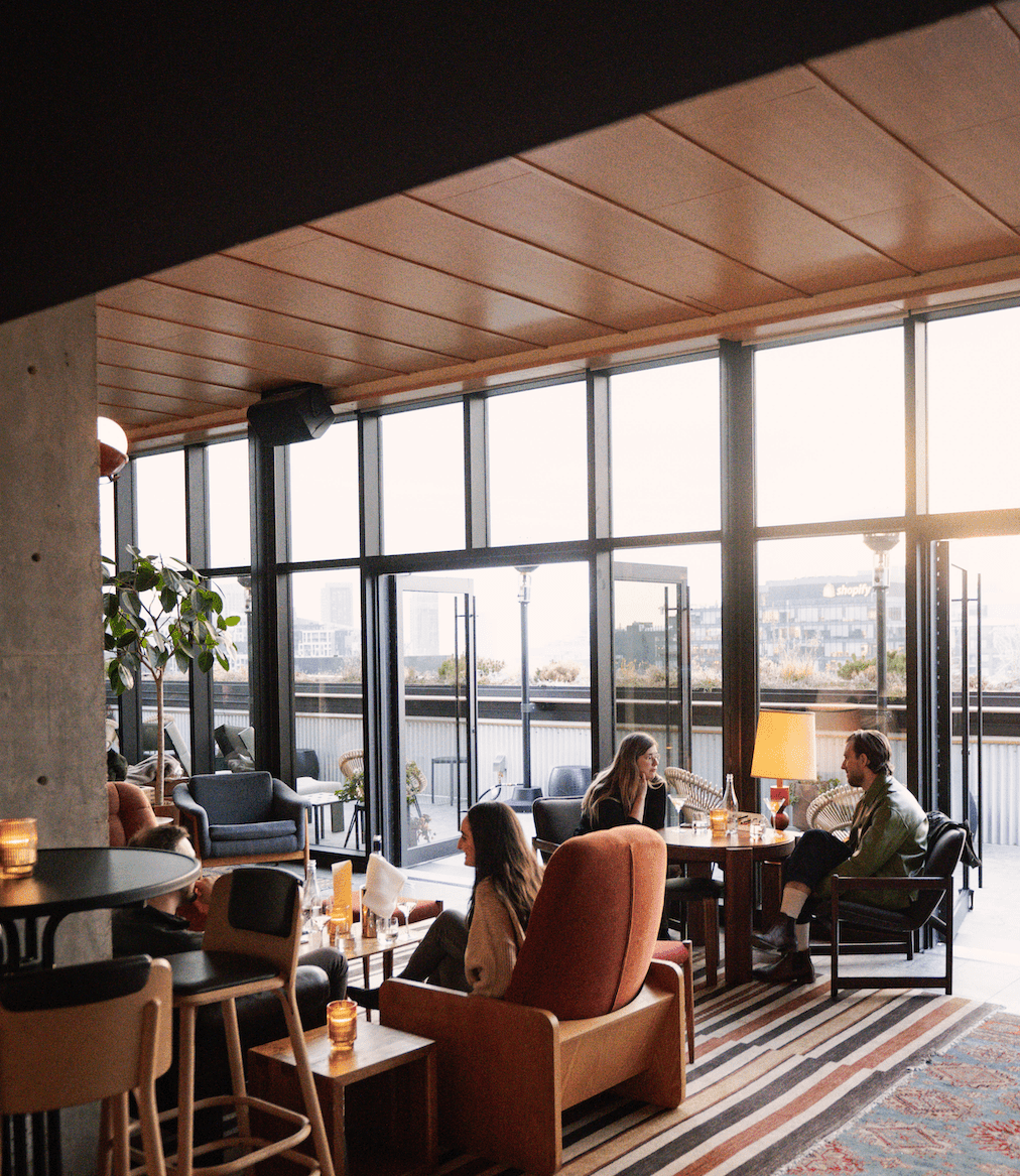 People sit and talk in a modern lounge with large windows, natural light, and city views. Candles are on the tables.